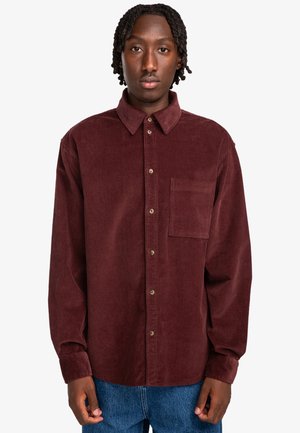 Chemise en velours côtelé bordeaux avec col boutonné, poche avant et boutons marron. Manches longues et coupe décontractée. Pas de motifs visibles.