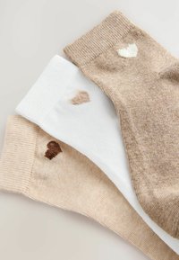 Tre par strumpor: beige, vit och ljusbeige. Varje par har ett litet hjärtmönster i kontrasterande färger vid mudden. Mjukt, texturerat tyg.