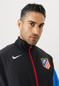 Must värviline spordijakk punase tõmbluku ja valge Nike logoga, millel on sinised aktsendid. Jakk sisaldab ka kilbi logo, millel on tähed ja triibud.