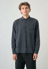 Schwarzes Langarmhemd aus weichem Stoff, mit einer durchgehenden Knopfleiste, klassischem Kragen und einer einzelnen Brusttasche. Glatte Textur.