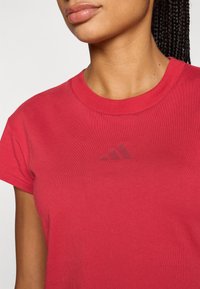 Κόκκινο βαμβακερό μπλουζάκι με κοντά μανίκια και στρογγυλή λαιμόκοψη. Διαθέτει τονικό λογότυπο Adidas στο στήθος. Λεία υφή κ�αι εφαρμοστό σχέδιο.