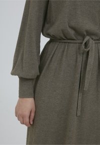 Vestito a coste di colore verde oliva con maniche lunghe e vita aderente con cintura annodata. Tessuto morbido con vestibilità rilassata e pieghe sottili.