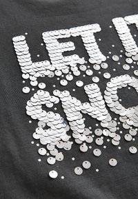 Grijze stof met zilveren pailletten die de tekst "LET IT SHINE" vormen, vergezeld van extra verspreide pailletten die textuur en glans creëren.