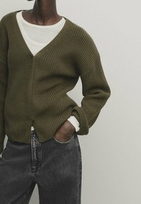 Persona che indossa un cardigan chunky di colore verde oliva sopra una maglietta a maniche lunghe bianca e jeans a vita alta grigio scuro, con una mano in tasca.