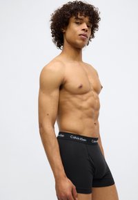 Boxeri scurți din bumbac negru, cu un brâu elastic moale purtând logo-ul "Calvin Klein". Modelul este fără tricou, arătând o fizionomie tonifiată.