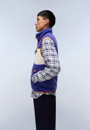 Fleece vest met een paars bovenstuk, een crèmekleurig onderpaneel en oranje accenten. Geruite shirt eronder, zwarte broek en details van zijzakken.