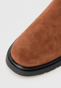 Botă din piele întoarsă maro, cu vârf rotund, având o talpă din cauciuc negru neted și o suprafață texturată confortabilă.