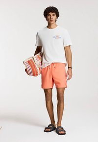 Bijela majica s plavim tekstom, koraljne kupaće hlače i tamne sandale. Drži prugastu torbu za plažu u crvenoj, narančastoj i krem boji.