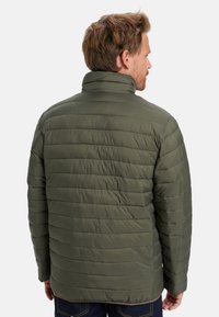 Hombre con chaqueta acolchada de color verde oliva y cuello alto, visto desde atrás, de pie contra un fondo claro.