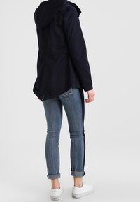 Granatowa kurtka z kapturem o luźnym kroju, z wciągniętym w talii i wentylacją na plecach. W połączeniu z podwiniętymi niebieskimi dżinsami i białymi sneakersami.