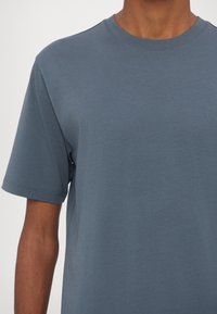 Gros plan d'une personne portant un t-shirt bleu uni à col rond et manches courtes, montrant le haut du torse et le bras droit.
