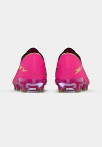 Vaaleanpunaiset jalkapallokengät, joissa on sulavalinjainen, teksturoitu päällinen, mustat yksityiskohdat ja läpinäkyvä pohja vihreillä nappuloilla, on suunniteltu suorituskykyä varten kentällä.