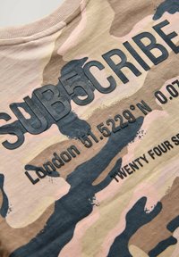 Krémszínű és rózsaszín árnyalatú kamuflázs póló, kiemelkedő fekete felirattal: "SUB5CRIBE," "London 51.5229° É 0.07°," és "TWENTY FOUR SEVEN."