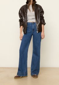 Žena v tmavě hnědé kožené bundě přes pruhované krátké top, s vysokým pasem, širokými modrými džíny a hnědými volnočasovými botami stojí uvnitř.