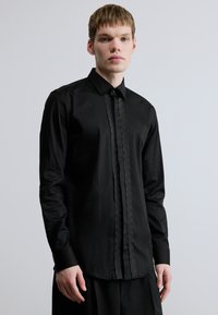 Fekete hosszú ujjú ing texturált gombolórésszel, egyenes gallérral és testhez simuló szabással. Sima anyag enyhe fényes felülettel.