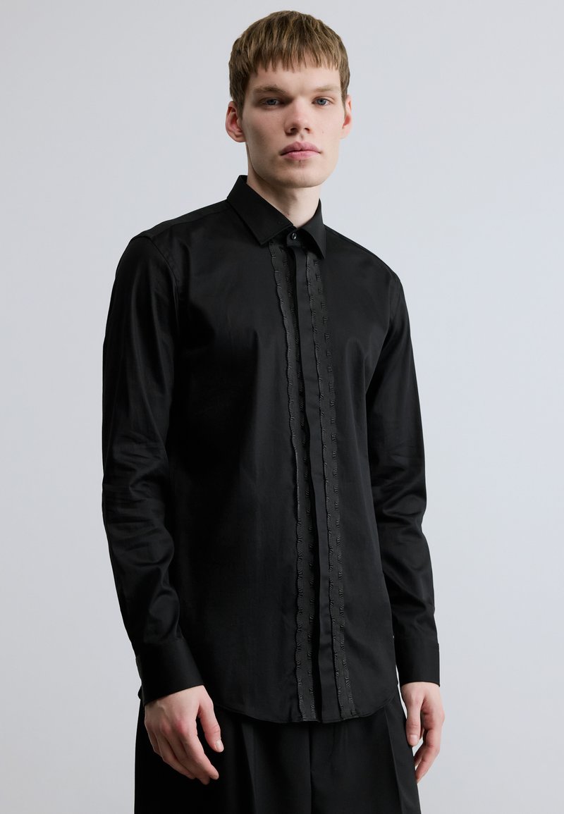 Fekete hosszú ujjú ing texturált gombolórésszel, egyenes gallérral és testhez simuló szabással. Sima anyag enyhe fényes felülettel.