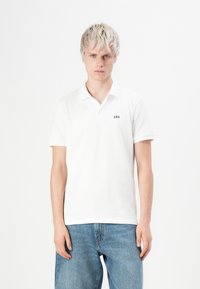 Jeune homme avec des cheveux blonds courts portant un polo blanc GAP et un jean bleu, se tenant devant un fond blanc uni.