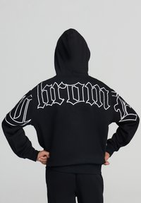 Persona che indossa un hoodie nero con il cappuccio sopra, con del testo in stile gotico bianco sulla schiena e sulle maniche, in piedi con le mani sui fianchi.