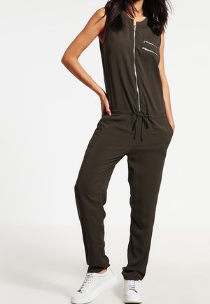 Ärmelloser olivgrüner Jumpsuit mit einem Reißverschluss vorne, Seitentaschen, verstellbarem Kordelzug in der Taille und kombiniert mit weißen Sneakers.