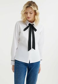 Camisa blanca de manga larga con una corbata negra en el cuello y detalles negros en los puños, hecha de una tela suave. Diseño de cuello clásico.