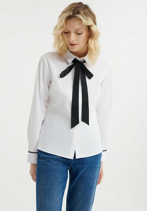 Camisa blanca de manga larga con una corbata negra en el cuello y detalles negros en los puños, hecha de una tela suave. Diseño de cuello clásico.