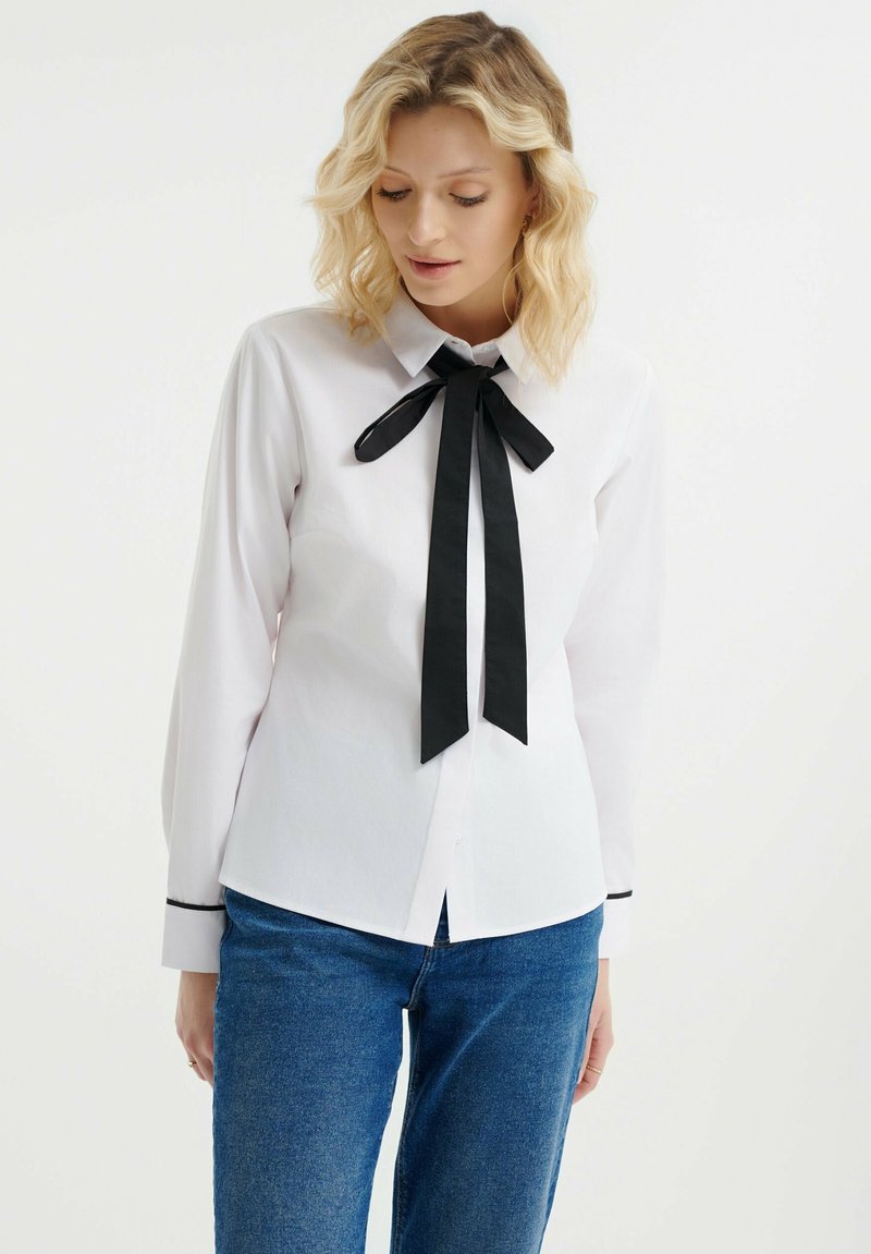 Camisa blanca de manga larga con una corbata negra en el cuello y detalles negros en los puños, hecha de una tela suave. Diseño de cuello clásico.