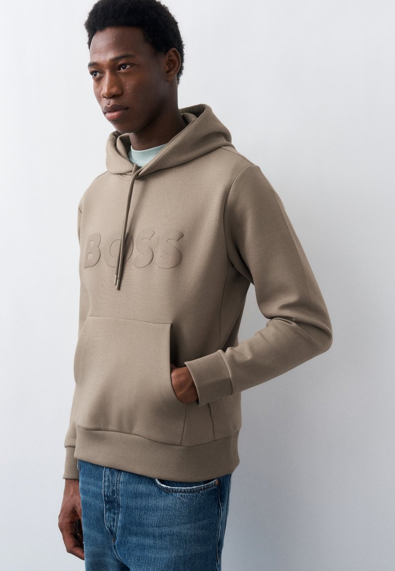 Νέος άνδρας που φοράει μπεζ φούτερ με ανάγλυφο κείμενο "BOSS" και μπλε τζιν, στέκεται με το ένα χέρι στην τσέπη απέναντι σε λευκό απλό φόντο.