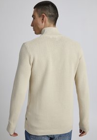 Cremfarbener, gerippter Strickpullover mit hohem Kragen, langen Ärmeln und taillierter Passform. Enthält strukturierte Details und ein kleines Logo-Element.