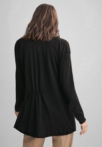 Svart långärmad topp med en samlad midjedetalj, mjuk textur och en lätt rundad kant, med raglanärmar för en avslappnad passform.