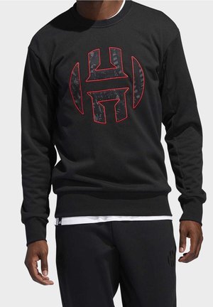 Man met een zwart sweatshirt met een groot gestileerd "H"-logo met een rode omlijning op de borst, gecombineerd met een zwarte broek en zichtbaar wit ondershirt.