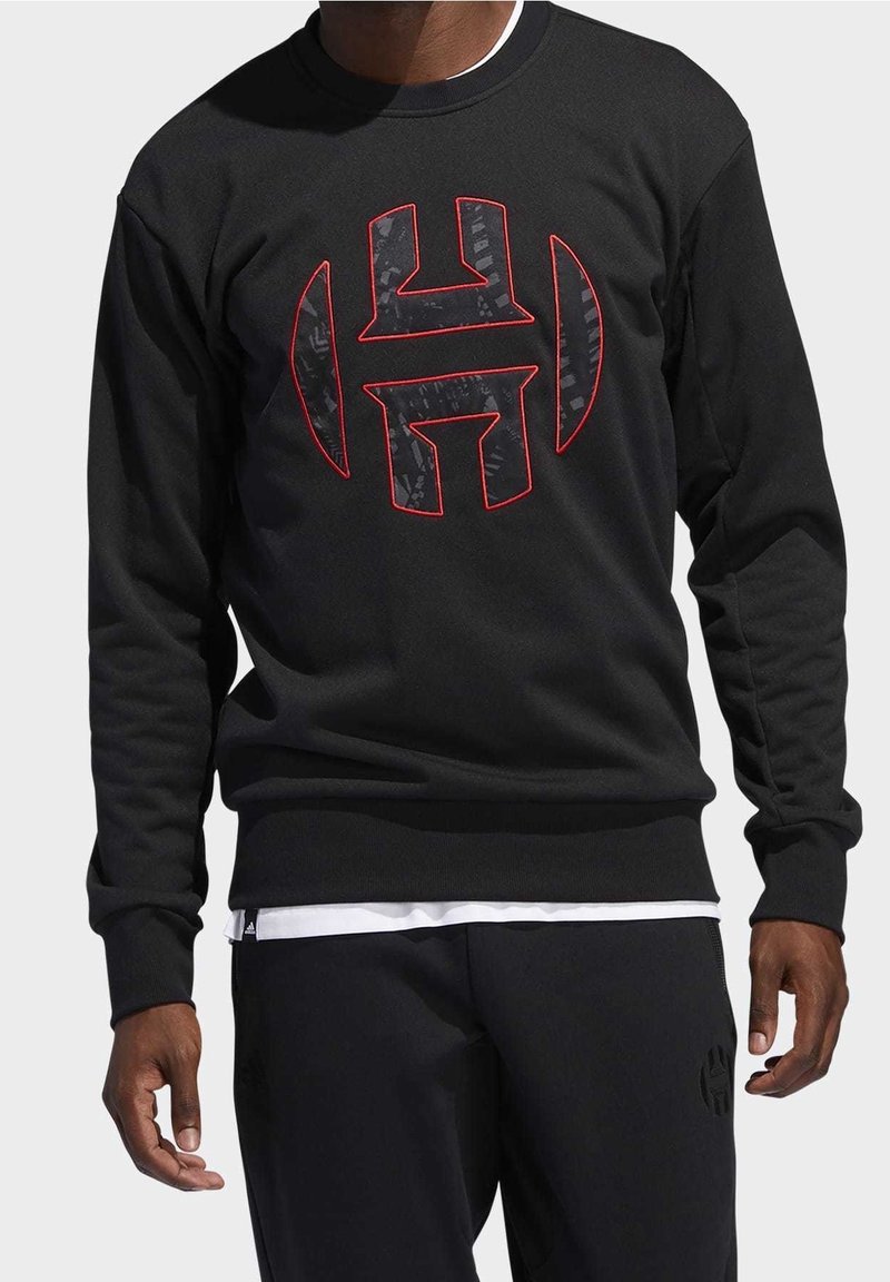 Homme portant un sweat-shirt noir avec un grand logo "H" stylisé et bordé de rouge sur la poitrine, assorti d'un pantalon noir et d'un sous-vêtement blanc visible.