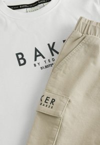 Valkoinen puuvillapaita, jossa on musta "BAKER by TED BAKER" -painatus, yhdistettynä beigekuituisiin housuihin, joissa on sivutasku ja etiketti.