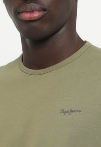 Oliverově zelené tričko s kulatým výstřihem, vyrobené z hladké tkaniny, se malým modrým logem "Pepe Jeans" na levé části hrudi.