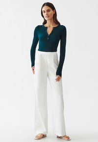 Teal ribbed long-sleeve top with button placket, paired with high-waisted white wide-leg trousers and minimalist sandals.