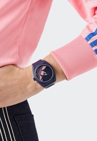 Ümar, tumesinine kell plastist rihmaga. Mustal kellakuval on roosad triibud ja näpunäidikud. Kandmiseks rannast randmel roosa varruka kõrval.