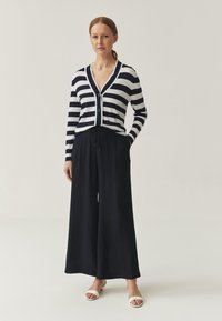 Cárdigan de botón a rayas en azul marino y blanco, combinado con pantalones negros de corte holgado y sandalias blancas. Diseño sencillo, ajuste casual.
