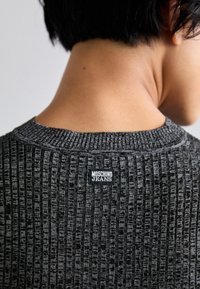 Pelēka ribota džemperis ar melnu etiķeti, uz kuras ir rakstīts "MOSCHINO JEANS", atpakaļ pie kakla, ar teksturētu adījuma apdari.