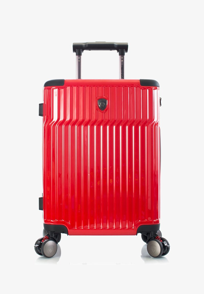 Valise rigide rouge avec poignée télescopique, rainures verticales texturées, quatre roues multidirectionnelles et logo central en forme de bouclier.