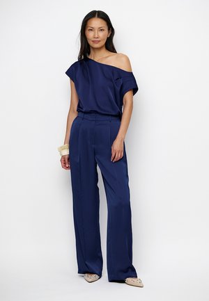 Vrouw draagt marineblauwe schouderloze blouse en wijde broekspijpen, beige puntige schoenen en witte armbanden aan de rechterpols, staand tegen een witte achtergrond.