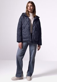 Giacca trapuntata blu navy con cappuccio, due tasche frontali e zip. Abbinata a jeans a gamba larga blu chiaro e sneakers crema.