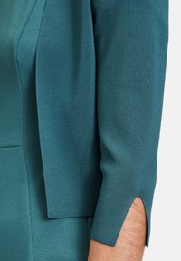 Turkusowa dzianinowa sukienka z materiału o ustrukturyzowanych rękawach z delikatnym wycięciem przy mankiecie. Gładka faktura i dopasowany krój.