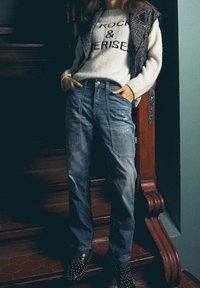 Mujer con un suéter blanco con texto, chaleco oscuro, jeans de tiro alto y deslavados, y zapatos negros con tachuelas, de pie en unas escaleras de madera.