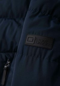 Sezione di giacca puffer blu navy con texture liscia, dotata di una spilletta in gomma nera con l'etichetta "team Ombre" vicino a una tasca con zip.