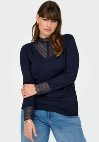 Dlouhý rukáv modrý top s krajkovou yoke a krajkovými detaily na manžetách, s ribovanou strukturou a zaobleným lem. Spárováno s modrými džíny.