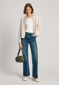 Veste bomber beige clair sur un débardeur blanc, associée à un jean bleu taille haute et des baskets crème. Tenant un petit sac à main vert.