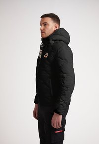Svart puffjacka med huva, med en framzipper och en logotyp patch på bröstet. Texturerad, quiltad tyg; slankt mönster.