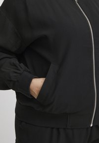 Čierna bombrová bunda s predným zipsom, rebrovanými manžetami a lemom, bočnými vreckami a hladkou textúrou látky.