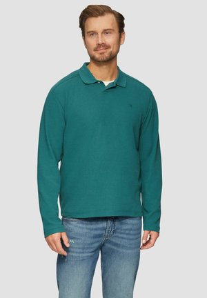 Polo à manches longues teal avec col, tissu texturé et un petit logo sur la poitrine gauche, associé à un jean bleu clair déchiré.
