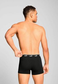 Man seen from the back adjusting black Puma boxer briefs with logo waistband, against plain light gray background.