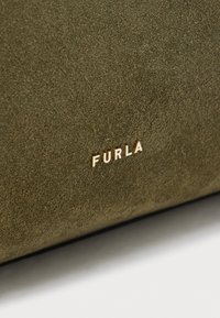 Olivzaļas zamšāda virsma ar nelieliem zelta krāsas "FURLA" logotipiem, kas iespiesti pie apakšējās malas.